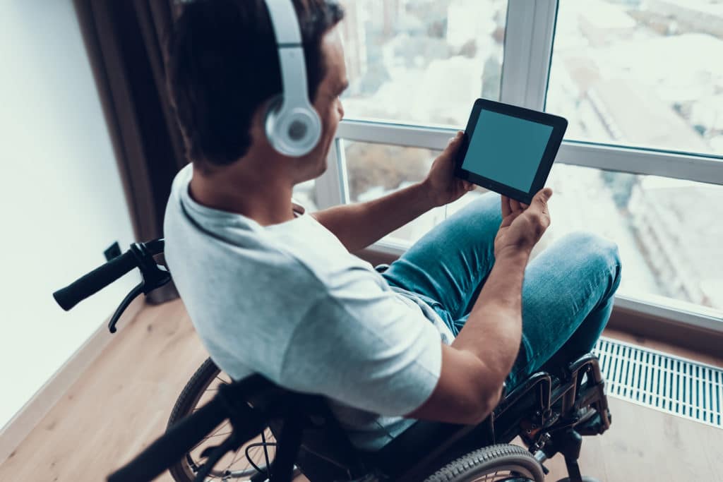 Improve Accessibility Back View Disabled Man in Headphones Uses Tablet. Closeup of Handsome Happy Person in Casual Clothes Sits in Modern Wheelchair Parked near Window in Living Room or Hospital. Man Watch at Empty Screen